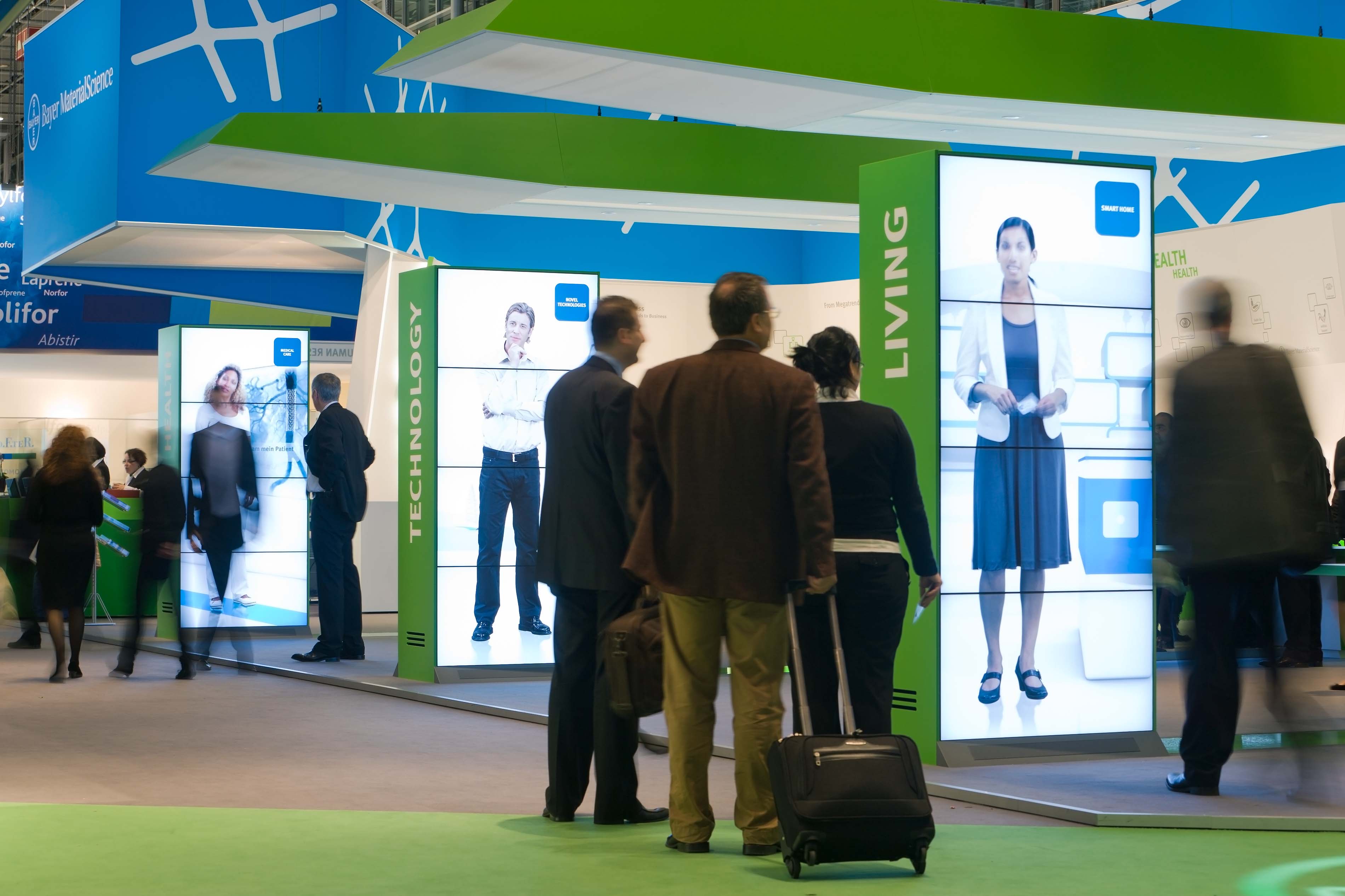 Trade fair visitors stand in front of the boothd. Information pillar shows film